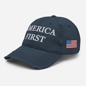 Puede incluir: Gorra de béisbol azul marino con las palabras "AMERICA FIRST" bordadas en blanco. Un parche de la bandera estadounidense está en el lateral. La gorra tiene un aspecto desgastado.