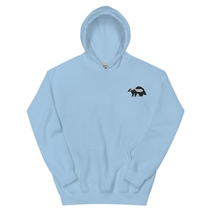 May include: A light blue hoodie with a kangaroo pocket and a drawstring hood. The hoodie features a small embroidered badger design on the upper left chest. The badger is black and white.