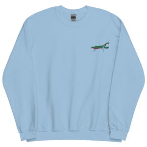 Pode incluir: Camisola azul clara com um pequeno desenho bordado de louva-a-deus em verde e vermelho no peito. A camisola tem gola redonda e mangas compridas. A etiqueta da marca é visível no interior da gola.