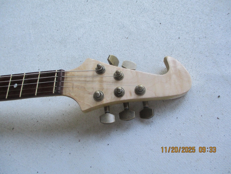 May include: Close-up of a guitar headstock with tuning pegs. The headstock is made of light-coloured wood with a curved design. The strings and fretboard are visible, along with the tuning pegs. The date and time are printed in the bottom right corner.