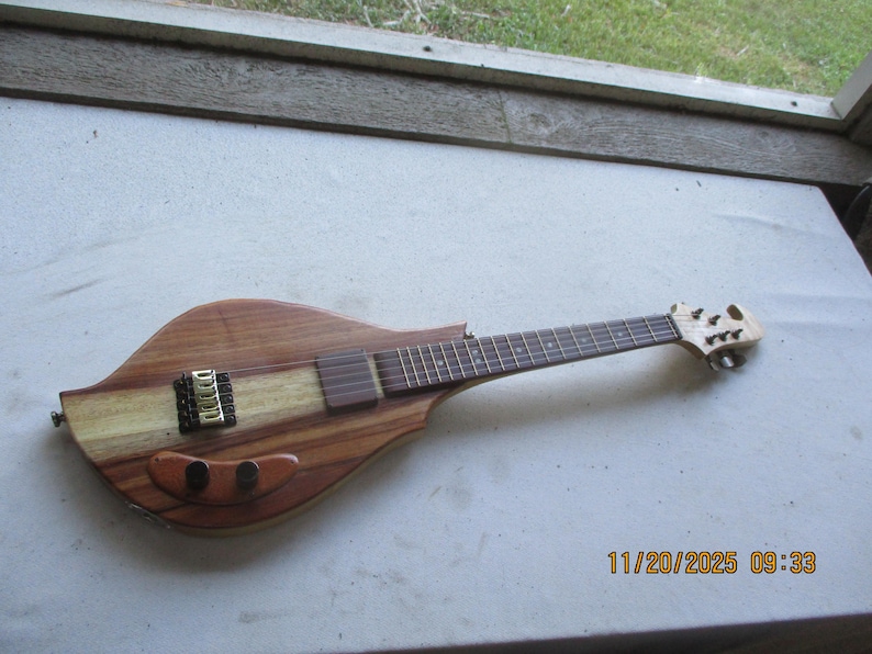 May include: A handcrafted electric guitar with a distinctive body shape. It has a light wood central section, a dark fretboard, and a gold-coloured bridge. The headstock is intricately designed. The guitar is made of wood.