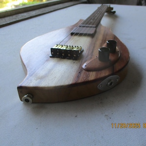 May include: A custom-made, teardrop-shaped electric guitar with a natural wood finish. The guitar features a light-coloured wood body with a darker wood accent, a gold-coloured bridge, black knobs, and a silver input jack. The fretboard is visible.