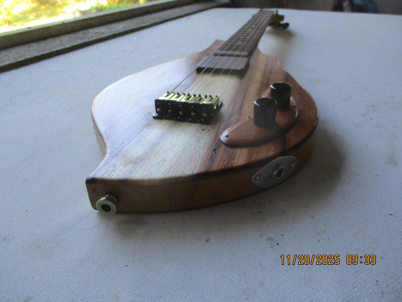 May include: A custom-made, teardrop-shaped electric guitar with a natural wood finish. The guitar features a light-coloured wood body with a darker wood accent, a gold-coloured bridge, black knobs, and a silver input jack. The fretboard is visible.