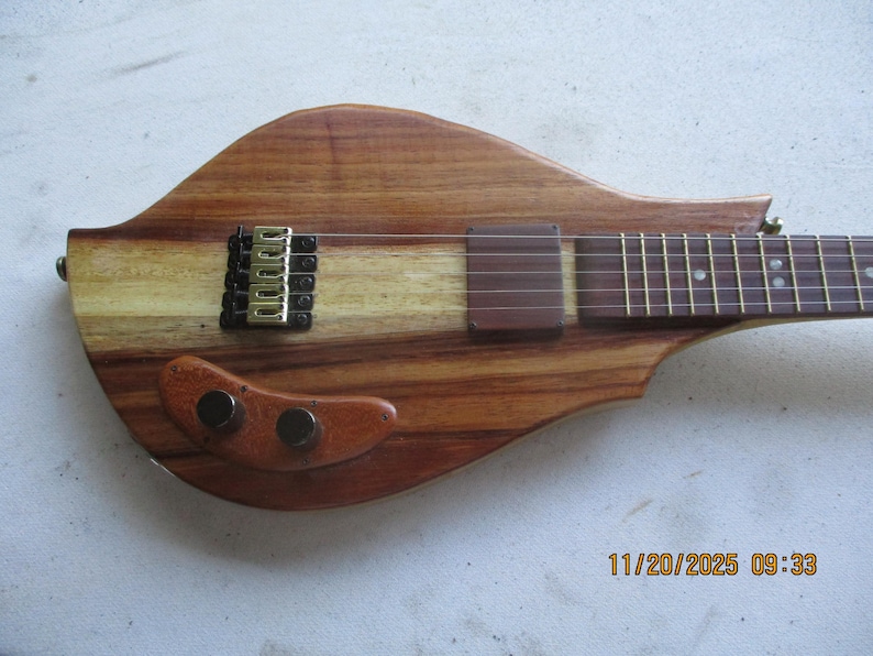 May include: A handcrafted wooden electric guitar with a distinctive, curved shape. The body is made from contrasting light and dark wood, with gold and black hardware. The fretboard has inlaid markers. The guitar's overall length is approximately 76 cm.