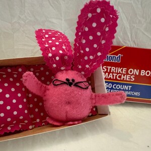 May include: A pink felt bunny with white polka dots, lying in a matchbox with a pink and white polka dot pillow. The matchbox is labeled "Diamond Strike on Box Matches 50 Count Kitchen Matches".