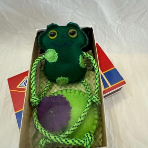 May include: A handmade green felt frog toy with yellow eyes and a green and yellow rope. The frog is in a box with a green lily pad with a purple flower. The box has red, blue, and yellow accents.