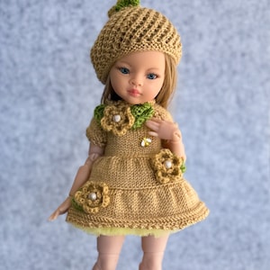 May include: A doll wearing a brown knitted dress with green and white flower accents, a brown knitted hat with green leaves, and brown shoes with bee accents.