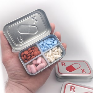 Puede incluir: Un pastillero de metal plateado abierto, que muestra compartimentos llenos de pastillas rojas, azules y blancas. La tapa tiene una "X" y una "B" estampadas. También se ve una segunda caja con un símbolo Rx rojo y blanco.