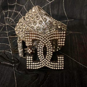 May include: Silver belt with a large buckle covered in sparkling rhinestones. The buckle features the letters "DG" and a star, also encrusted with rhinestones. The belt is on a black background with a spiderweb design.