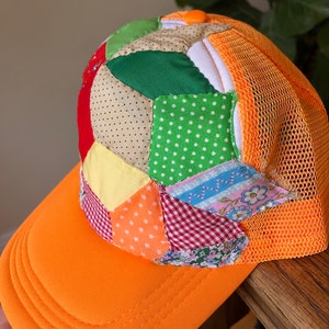 Puede incluir: Una gorra de béisbol naranja brillante con un diseño de patchwork en la corona. El patchwork está hecho de varias telas en diferentes colores y patrones, incluyendo verde, rojo, amarillo, azul y blanco. La parte posterior de la gorra está hecha de malla naranja.