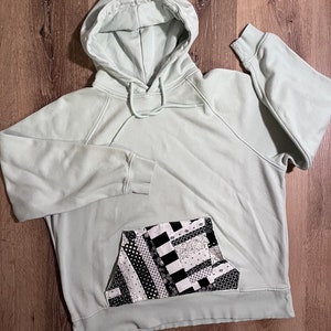 Puede incluir: Una sudadera con capucha verde claro con un bolsillo de patchwork blanco y negro.