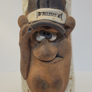 May include: A handcrafted ceramic mug with a whimsical face design. The mug features a brown, textured surface with a sculpted face, including a hat with the word "ALLSTAR". The mug has a beige and brown color scheme.