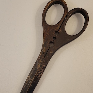 May include: Vintage, dark brown scissors with large finger holes. The scissors have a long, pointed blade and a riveted joint. The metal shows signs of wear and age, with a distressed finish. The scissors are likely used for cutting fabric or paper.