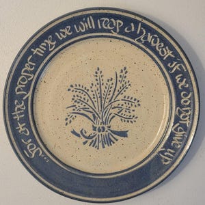 May include: A ceramic plate with a blue rim and a cream-colored center. The plate features a blue wheat sheaf design and the text "For at the proper time we will reap a harvest if we do not give up."