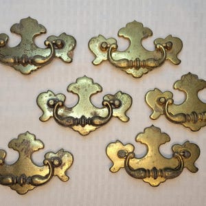 May include: A set of six brass drawer pulls, each featuring a decorative backplate with a curved, swinging handle. The metal has a tarnished, aged finish with visible patina and intricate, symmetrical scrollwork details on the mounting plates.