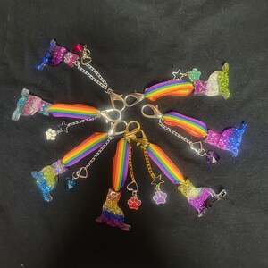 May include: A set of seven rainbow-colored keychains featuring a cat design. Each keychain has a different color combination of rainbow stripes and a small charm attached. The charms include a star, a paw print, and a heart.