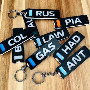 Könnte beinhalten: Eine Sammlung schwarzer, rechteckiger Schlüsselanhänger mit weißer Schrift und farbigen Akzenten. Jeder Schlüsselanhänger hat einen silbernen Schlüsselring. Die Schlüsselanhänger liegen auf einer Holzoberfläche verstreut, mit verschiedenen Wörtern.