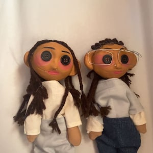 May include: Two handmade dolls with button eyes, rosy cheeks, and brown hair. One doll wears a white shirt and light blue pants, the other wears a white shirt and blue jeans. Both dolls have a unique, whimsical design.