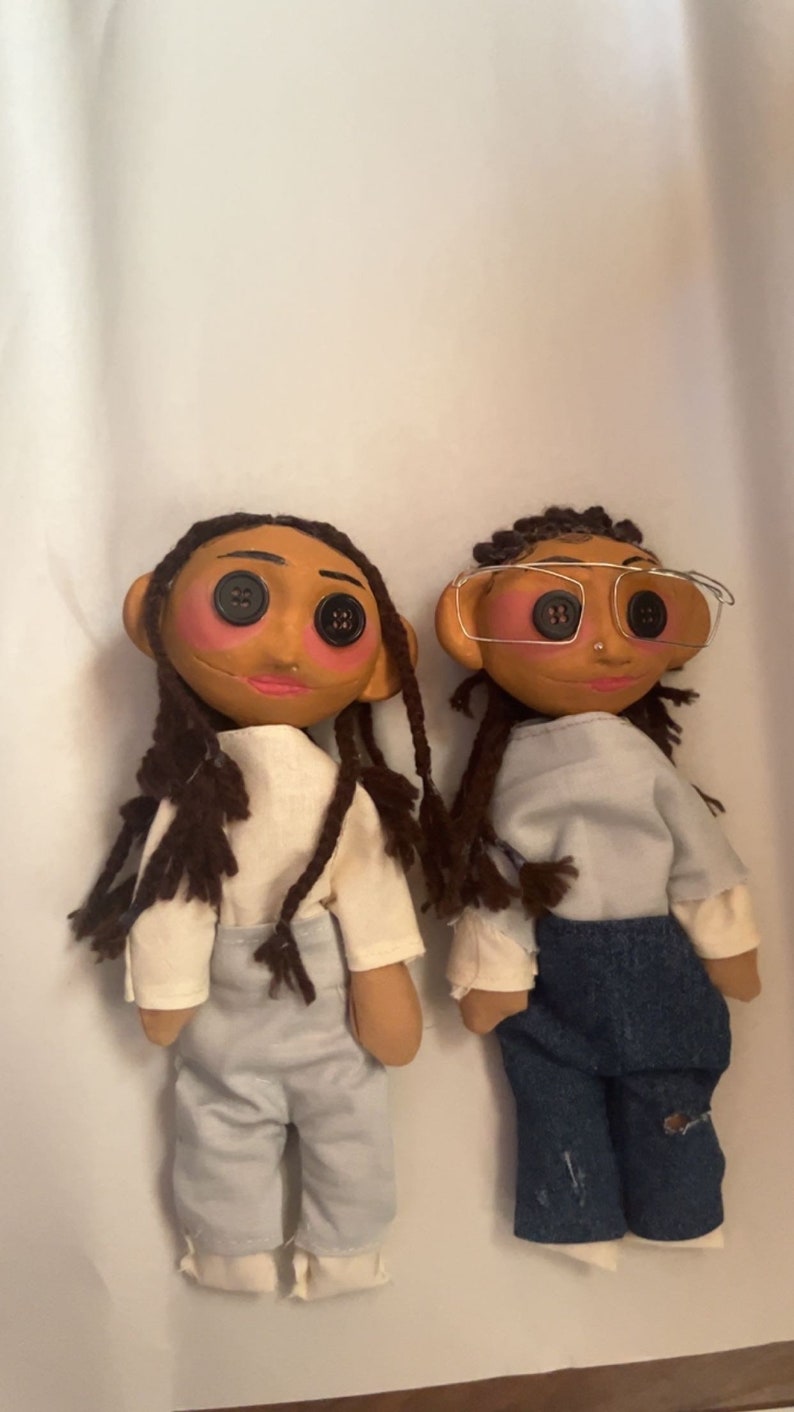 May include: Two handmade dolls with button eyes, rosy cheeks, and brown hair. One doll wears a white shirt and light blue pants, the other wears a white shirt and blue jeans. Both dolls have a unique, whimsical design.