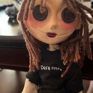 May include: A handmade doll with button eyes, brown yarn hair, and a black t-shirt with the text "Deft ones." The doll has a pale face with rosy cheeks and dark red lipstick, and is wearing blue denim jeans.