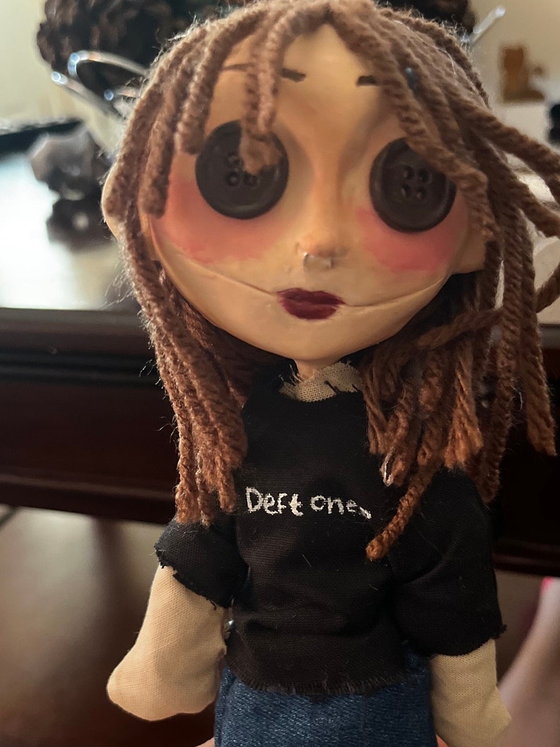 May include: A handmade doll with button eyes, brown yarn hair, and a black t-shirt with the text "Deft ones." The doll has a pale face with rosy cheeks and dark red lipstick, and is wearing blue denim jeans.