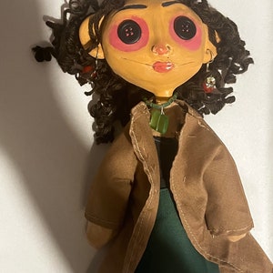 May include: A handmade doll with a tan face, button eyes, and curly brown hair. The doll wears a brown coat over a green top and a green pendant necklace. Earrings and a painted face complete the look.