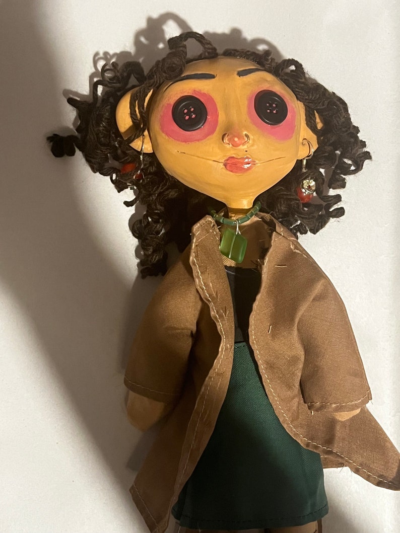 May include: A handmade doll with a tan face, button eyes, and curly brown hair. The doll wears a brown coat over a green top and a green pendant necklace. Earrings and a painted face complete the look.