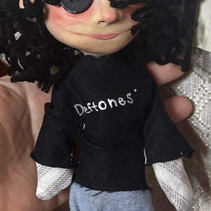May include: A handmade doll with button eyes, black curly hair, and a black shirt with the band name "Deftones" in white. The doll is wearing blue jeans and has a painted face.