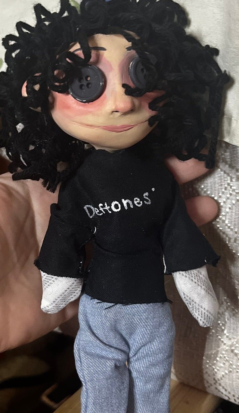 May include: A handmade doll with button eyes, black curly hair, and a black shirt with the band name "Deftones" in white. The doll is wearing blue jeans and has a painted face.