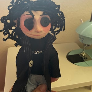 May include: Handmade doll with button eyes and black curly hair. The doll wears a black coat, a black shirt with the word "honest" printed on it, and blue jeans. The doll's face is pale with rosy cheeks.