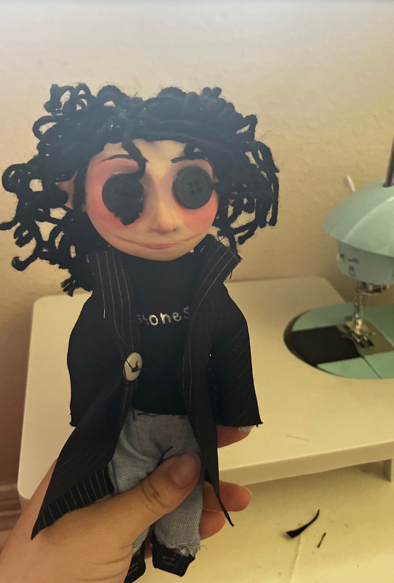 May include: Handmade doll with button eyes and black curly hair. The doll wears a black coat, a black shirt with the word "honest" printed on it, and blue jeans. The doll's face is pale with rosy cheeks.
