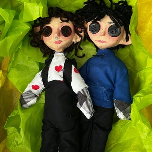May include: Two handmade dolls with button eyes and yarn hair. One doll wears a white shirt with red heart accents and black overalls. The other doll is dressed in a blue shirt and black pants. They are nestled in a bed of green tissue paper.