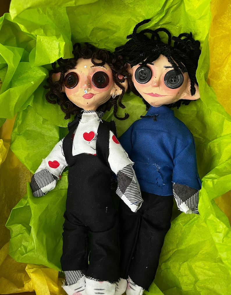 May include: Two handmade dolls with button eyes and yarn hair. One doll wears a white shirt with red heart accents and black overalls. The other doll is dressed in a blue shirt and black pants. They are nestled in a bed of green tissue paper.