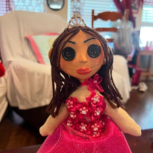 May include: A handmade doll with button eyes, brown hair, and a tiara. The doll wears a pink dress with floral embellishments and a matching choker. The doll's face is painted with red lips and has a neutral skin tone.