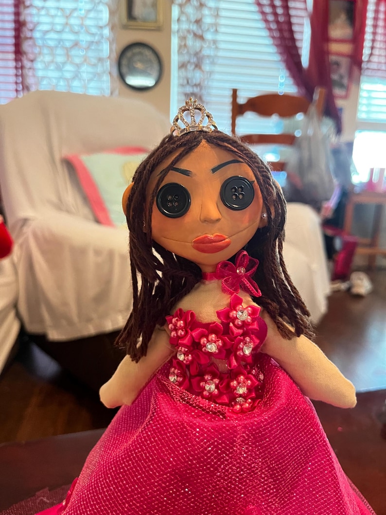 May include: A handmade doll with button eyes, brown hair, and a tiara. The doll wears a pink dress with floral embellishments and a matching choker. The doll's face is painted with red lips and has a neutral skin tone.