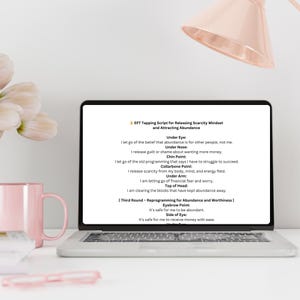 K&ouml;nnte beinhalten: Ein Laptop zeigt ein EFT-Tapping-Skript zur Freisetzung von Mangeldenken und zur Anziehung von F&uuml;lle. Der Bildschirm zeigt Text mit Affirmationen. Eine rosa Tasse und ein Stift liegen auf dem wei&szlig;en Schreibtisch.