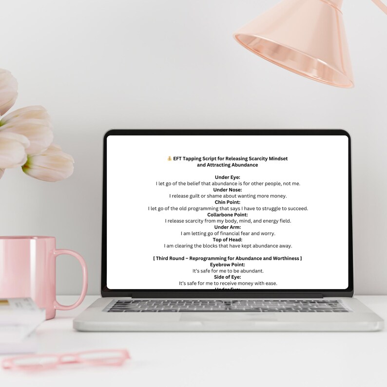 K&ouml;nnte beinhalten: Ein Laptop zeigt ein EFT-Tapping-Skript zur Freisetzung von Mangeldenken und zur Anziehung von F&uuml;lle. Der Bildschirm zeigt Text mit Affirmationen. Eine rosa Tasse und ein Stift liegen auf dem wei&szlig;en Schreibtisch.