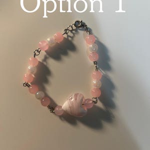May include: A bracelet featuring pink and white beads with a central heart-shaped bead. The bracelet includes a silver-toned metal chain and a lobster claw clasp. The text 'Option 1' is visible at the top of the image, indicating a selection choice.