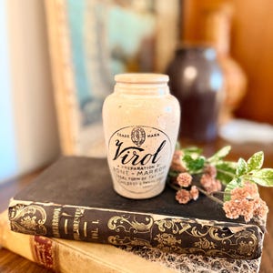 May include: A vintage cream-colored ceramic jar with a narrow neck and a wide base. The front features black printed text that reads Virol, Trade Mark, A Preparation of Bone Marrow, Ideal Form of Fat for Children and Invalids.