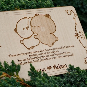 May include: A wooden plaque featuring a laser-etched design of two hugging cartoon bears. The plaque includes a loving message and the names "Anna" and "Adam" engraved below. Decorative floral elements adorn the edges.