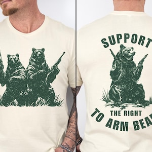 May include: A beige t-shirt with a green illustration of two bears holding guns. The back of the shirt has the text "SUPPORT THE RIGHT TO ARM BEARS".