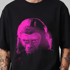 Könnte beinhalten: Ein schwarzes Kurzarm-T-Shirt mit einem leuchtend pinkfarbenen Grafikdruck eines Primatengesichts auf der Brust. Das Design zeigt eine detaillierte, kontrastreiche Illustration eines Schimpansen mit intensivem Blick, mittig auf der Vorderseite.