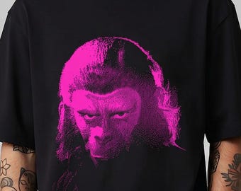May include: A black short-sleeve t-shirt featuring a vibrant hot pink graphic print of a primate face on the chest. The design displays a detailed, high-contrast illustration of a chimpanzee with intense eyes, centered on the front of the garment.