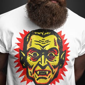 May include: White t-shirt featuring a vintage-style illustration of a vampire's face in yellow and black, with a red burst background. The t-shirt is worn by a person with a long beard and sunglasses.