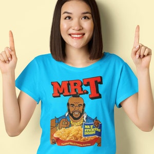 May include: A light blue t-shirt featuring a graphic of Mr. T holding a bowl of cereal. The text "MR.T" is in red block letters above the graphic. The cereal box reads "Mr. T Stickers Inside" and "Flavored With B-Vitamins And Iron."