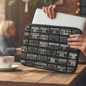 Könnte beinhalten: Eine rechteckige Laptoptasche mit einem sich wiederholenden Muster aus verschiedenen Vintage-Boombox-Kassettenrekordern in Schwarz-, Grau- und Silbertönen. Die Tasche hat einen schwarzen Rand und enthält einen dünnen, silbernen Laptop.