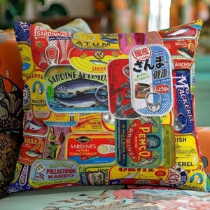 May include: Square throw pillow with a collage of vintage sardine can labels in various colors. The labels feature text in multiple languages, illustrations of fish, and brand names. The pillow is on a floral patterned chair.