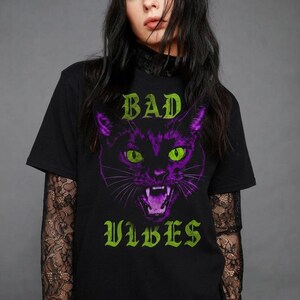 May include: A black short-sleeved t-shirt featuring a graphic of a snarling cat with purple fur and bright green eyes. The text "BAD" appears in green above the cat, and the word "VIBES" appears in green below the cat, all set against a black background.