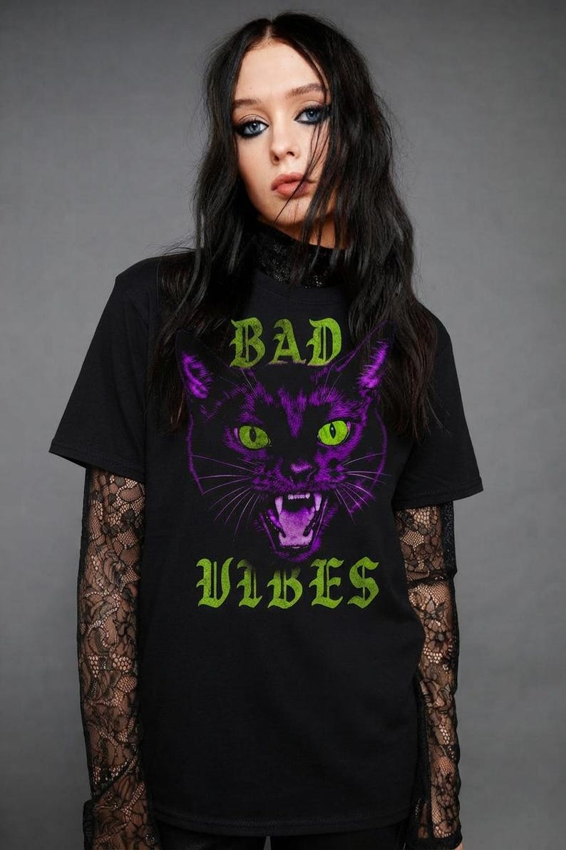May include: A black short-sleeved t-shirt featuring a graphic of a snarling cat with purple fur and bright green eyes. The text "BAD" appears in green above the cat, and the word "VIBES" appears in green below the cat, all set against a black background.