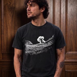 May include: A charcoal grey short-sleeve t-shirt featuring a graphic print of a small white fluffy dog sitting on a textured, rocky landscape. The shirt has a crew neckline and a faded, vintage-inspired wash throughout the fabric.